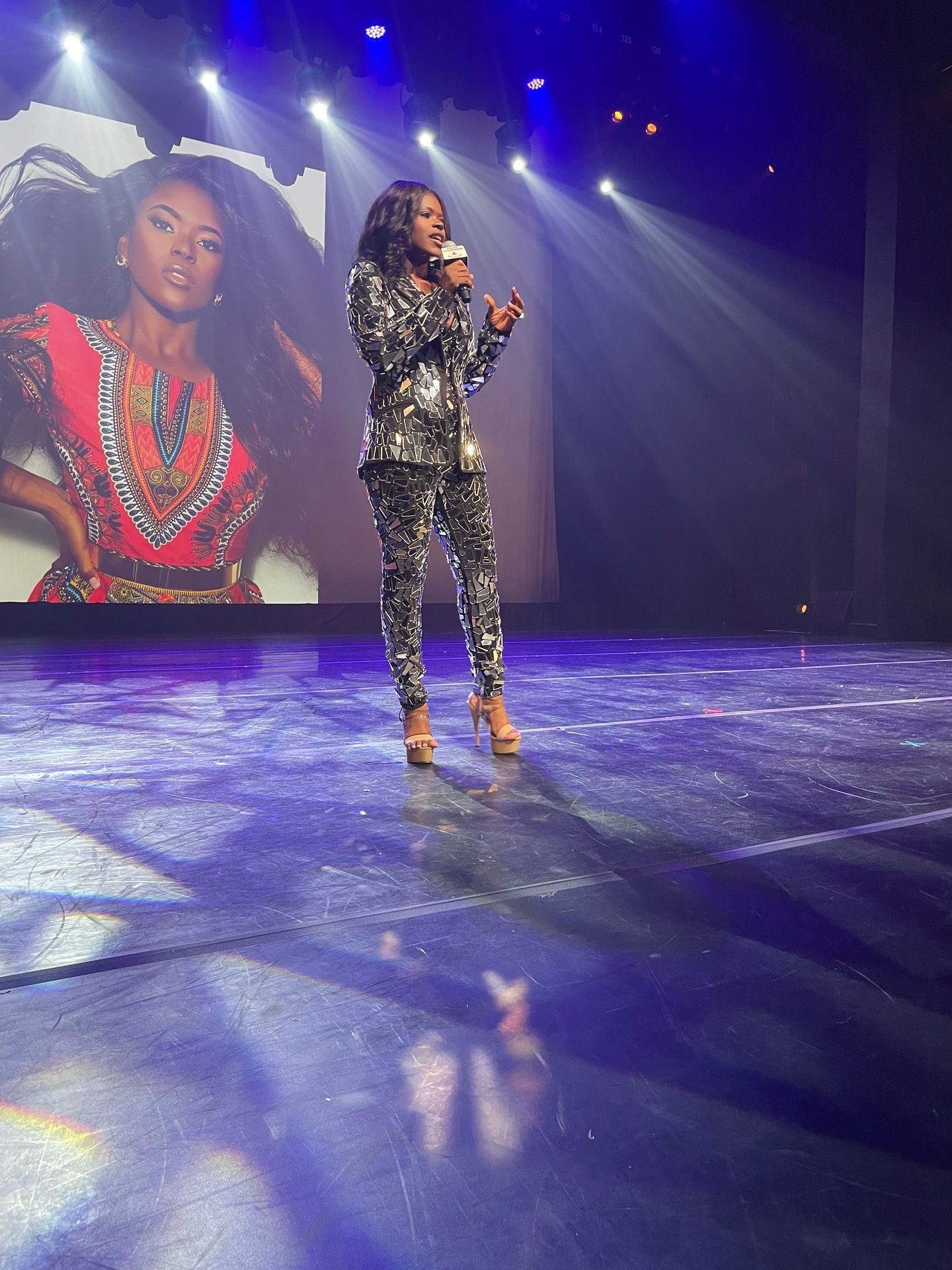 Person in patterned suit and heels speaking on stage with a large screen displaying an image behind them.